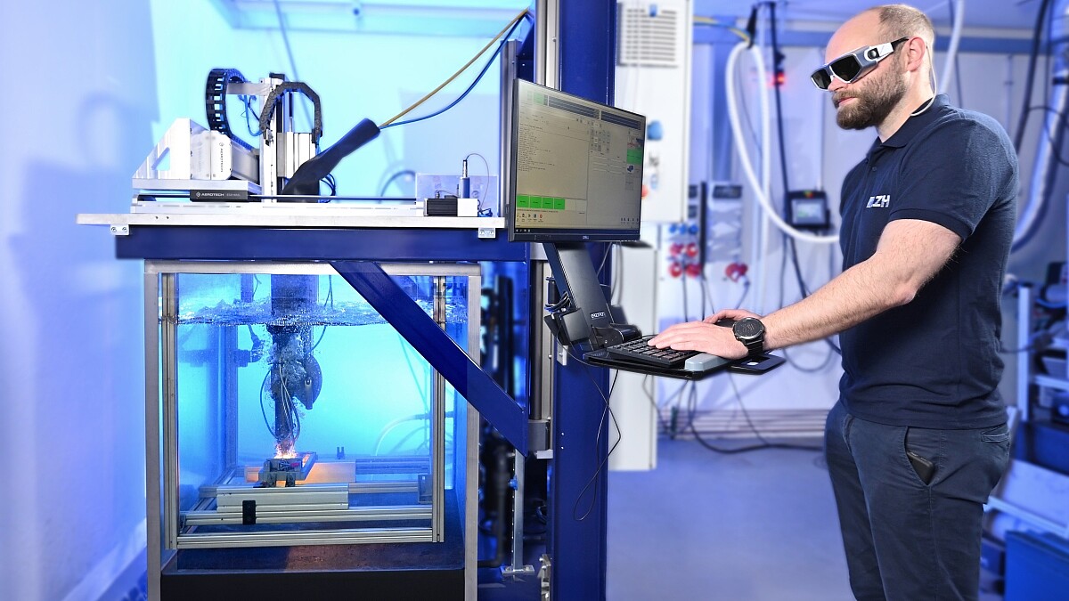 Ein Wissenschaftler mit Schutzbrille steht vor einem Wassertank, in dem ein Laser arbeitet, und bedient eine Tastatur unter einem Display.