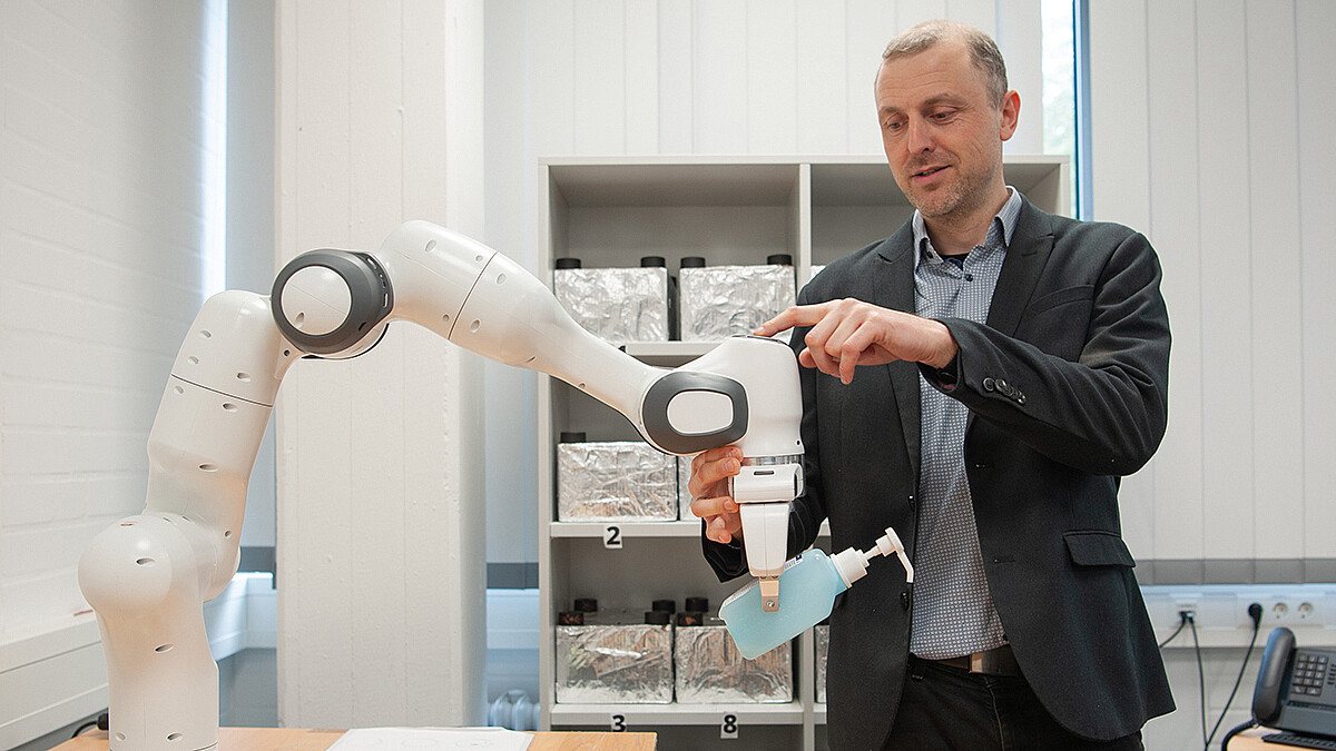 Der Forscher berührt mit seinen Händen ein Gelenk des Roboterarms, der eine Dosierflasche hält.