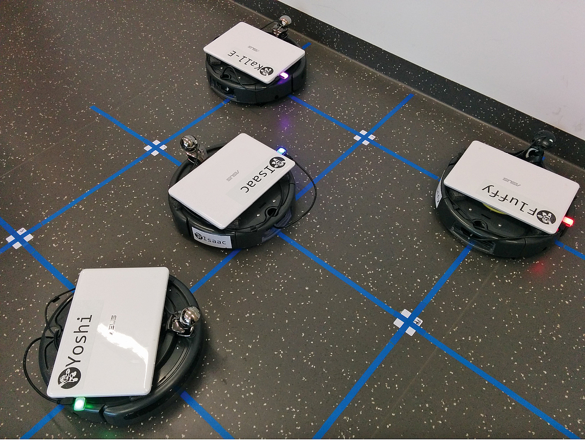Vier tellerförmige Roboter mit Laptop fahren Gitterlinien auf dem Boden ab.