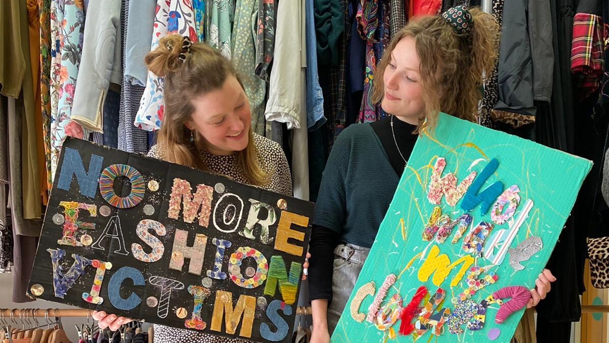 Zwei Studentinnen stehen vor Second-Hand-Kleidung und zeigen Plakate mit den Slogans „No more fashion victims“ und „Who made my clothes?“.