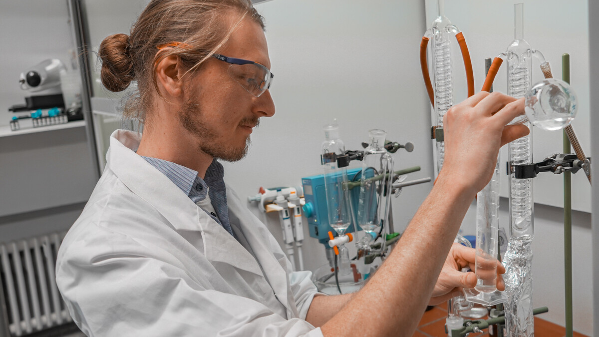 Ein junger Mann mit Laborkittel und Schutzbrille steht in einem chemischen Labor und füllt eine klare Flüssigkeit aus einem Kolben in einen Standzylinder.