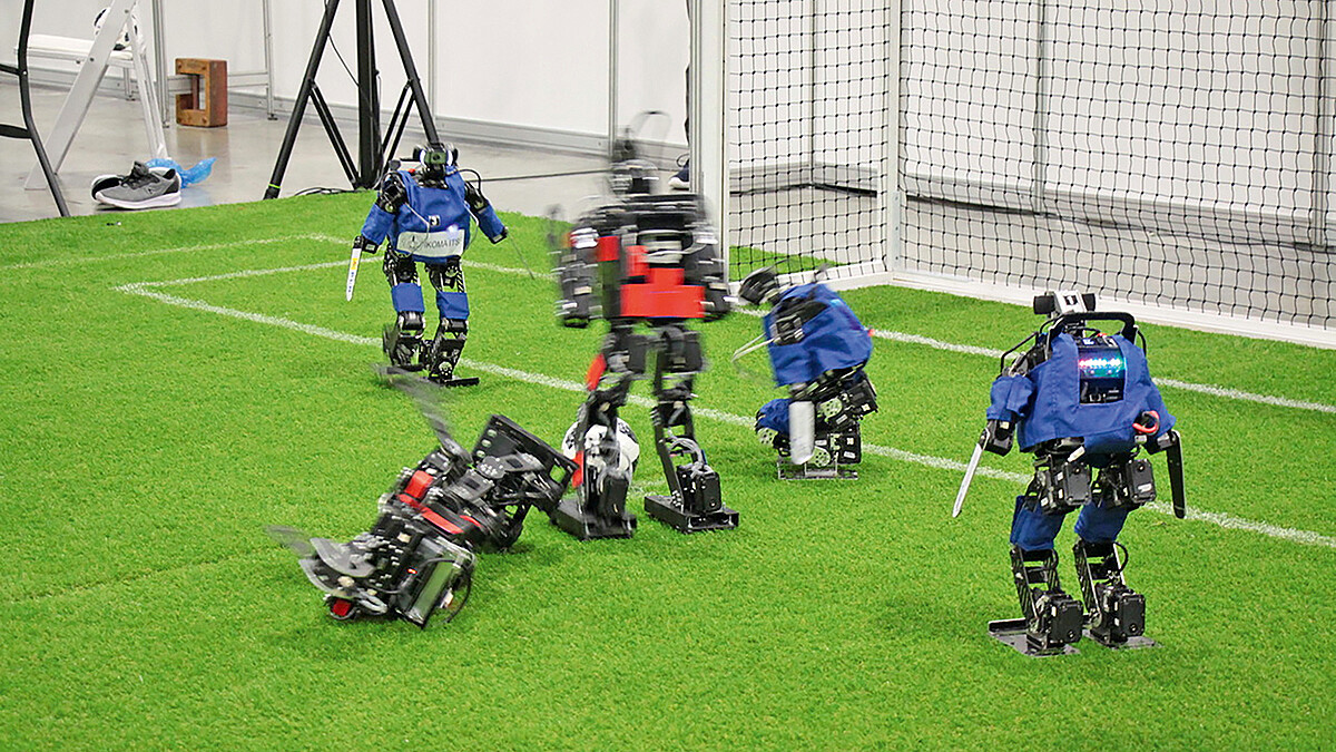 Fünf humanoide Roboter in Modellgröße, die farblich zwei Teams zugeordnet sind, stehen auf Kunstrasen vor einem Fußballtor. Ein Roboter liegt auf der Seite.