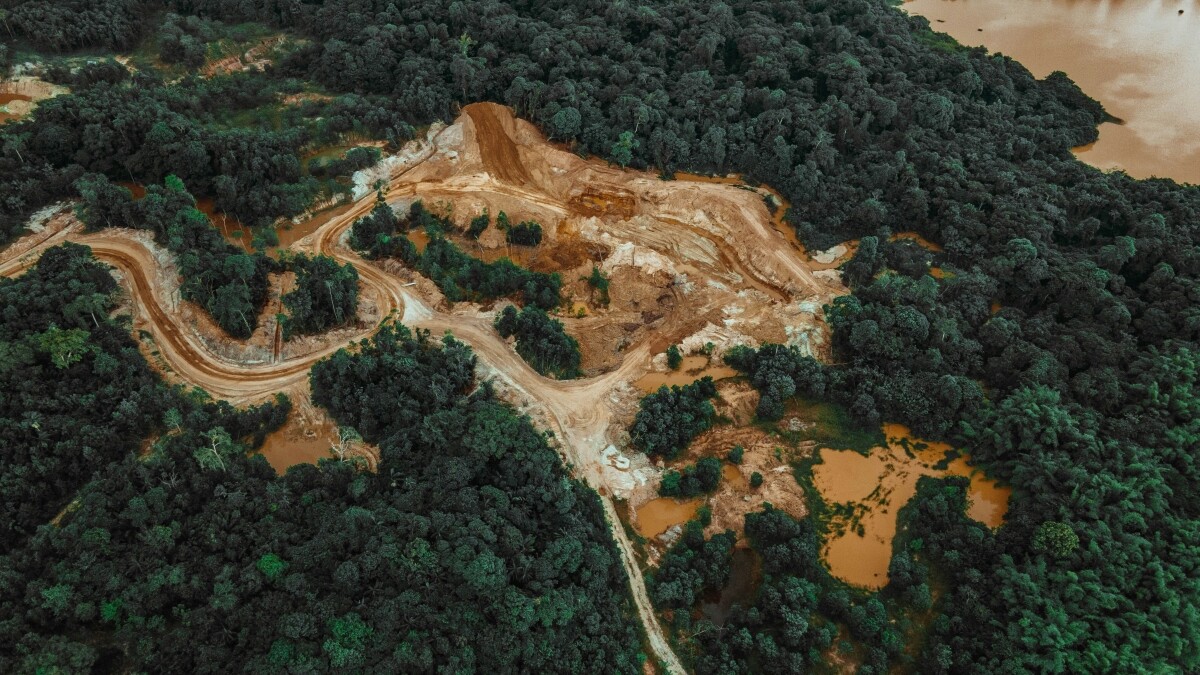 Die Luftaufnahme zeigt einen tropischen Wald. Mitten zwischen den grünen Bäumen erstreckt sich ein großer baumfreier Bereich mit Zufahrten, nackter Erde, Steinen und Tümpeln mit braunem Wasser.
