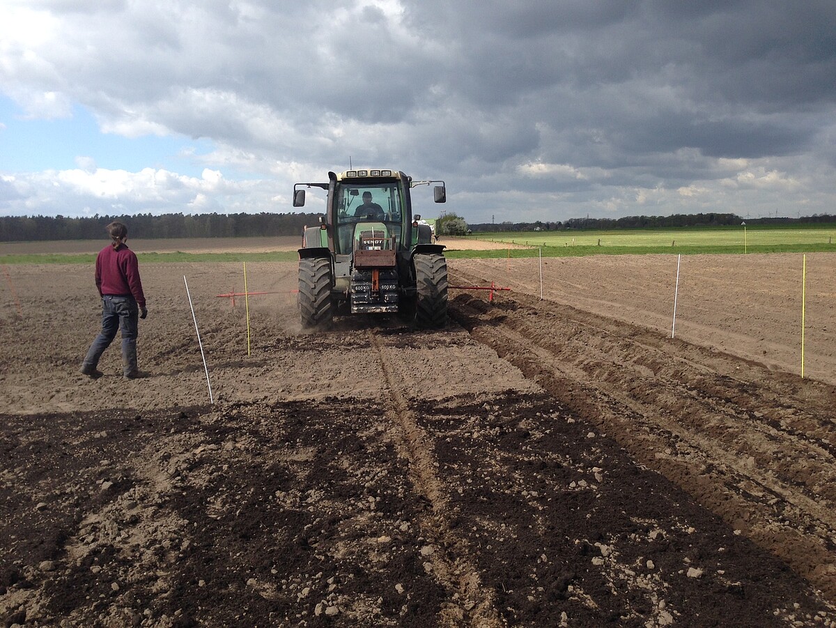 Neben einer dunklen, kompostierten Parzelle fährt ein Traktor über ein helleres, ungedüngtes Feld. Eine weitere Person schaut zu.