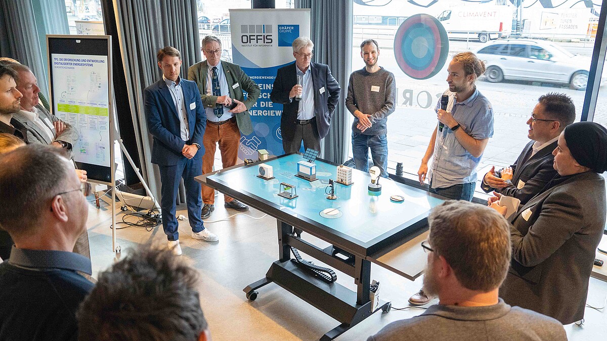 Eine Gruppe von Personen steht um einen Arbeitstisch, auf dem handgroße Modelle wie Gebäude, Solarspeicher und Glühlampe stehen.