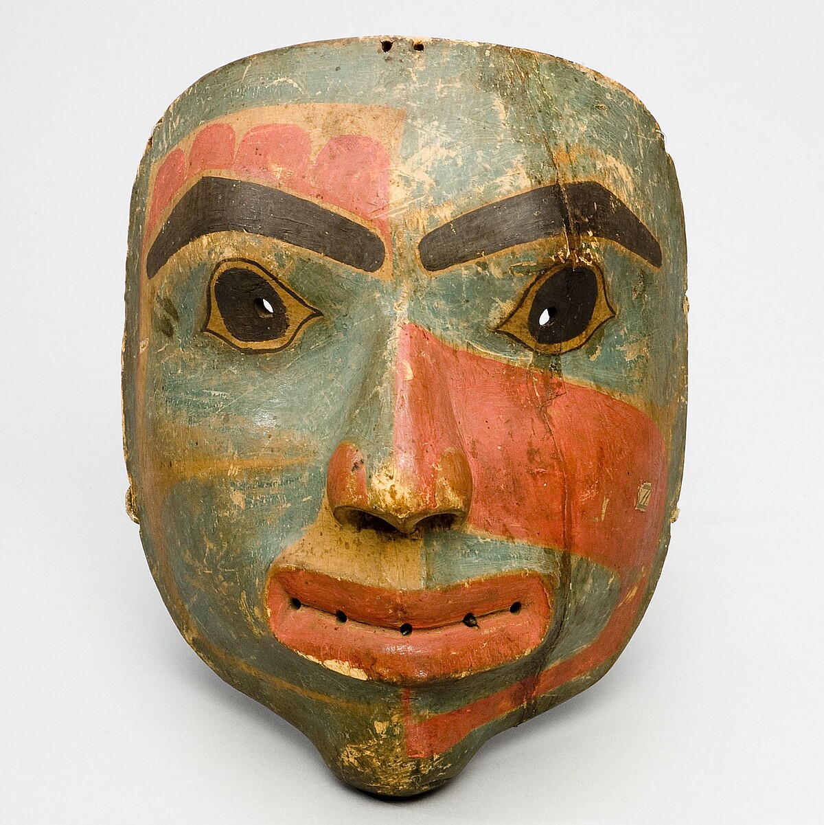 Die Maske hat die Form eines Gesichts, zeigt große Augen, prägnante Ausgenbrauen und Lippen sowie eine dreifarbige Gesichtsbemalung mit Formen und Mustern.