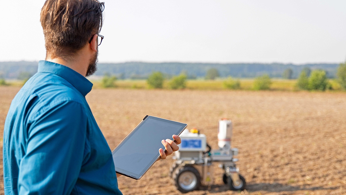 Eine Person hält ein Tablet in der Hand fest. Im Hintergrund steht der Agrarroboter auf der Ackerfläche.