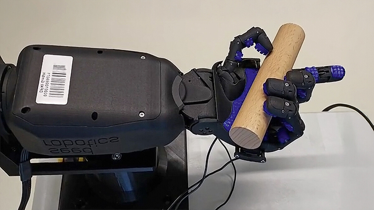 Eine Roboterhand mit Noppen an Gelenken und Gliedern hält mit Daumen und drei Fingern einen kurzen Holzzylinder. Der Zeigefinger ist ausgetreckt.