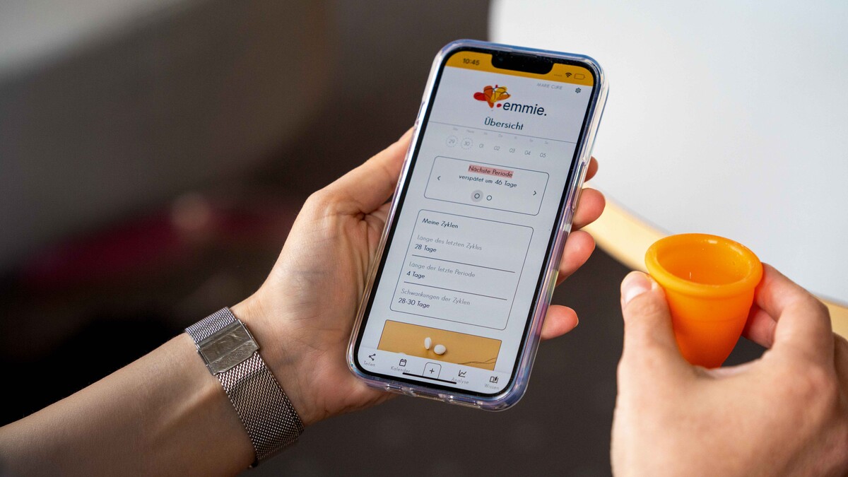 Eine Hand hält eine Menstruationstasse, die andere Hand hält ein Smartphone. Das Display zeigt Angaben zum Menstruationszyklus an.