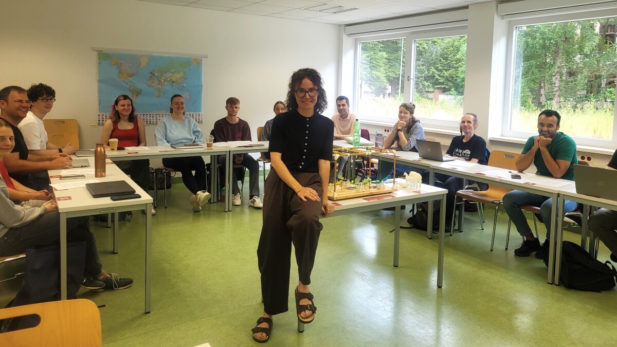 Heller Seminarraum mit U-förmig angeordneten Tischen. In der Mitte lehnt eine Frau mit dunklen Locken, Brille, schwarzem Oberteil und brauner weiter Hose an einem Tisch und lächelt in die Kamera. Neben ihr steht ein Holzmodell mit farbigen Kunststoffhebeln und kleinen weißen Tierfiguren. Um sie herum sitzen rund ein Dutzend Personen an den Tischen.