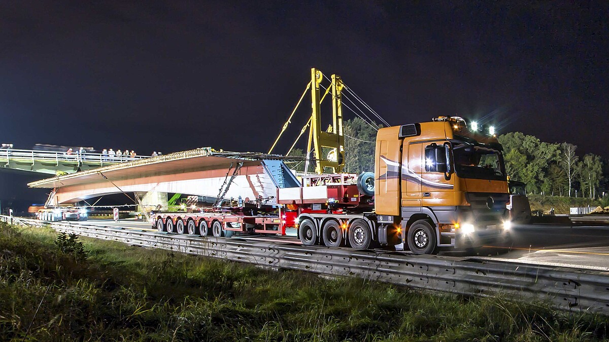 Ein Schwerlasttransporter mit einem Brückenbauteil fährt über eine Autobahnbaustelle.