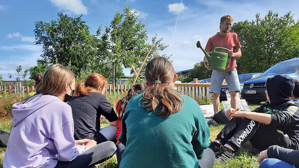 Kinder sitzen im Gras vor Obstbäumen. Eine Person mit Gießkanne in der Hand spricht zu ihnen.