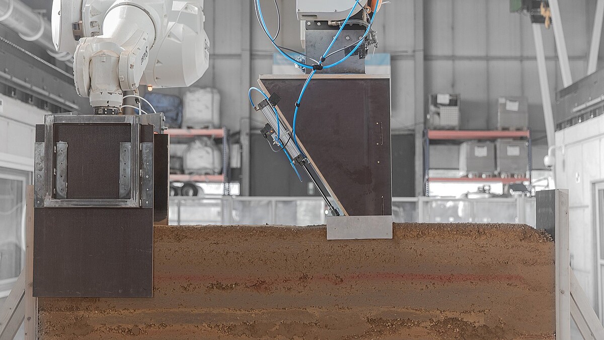 Ein Roboterarm platziert eine aktive Gleitschalung an der Lehmwand dort, wo verdichtet wird. Aus einem zweiten Roboterarm wird Lehm aufgetragen.
