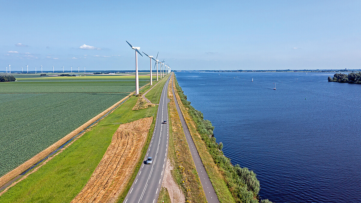 Autos fahren auf einer Landstraße am Wasser an Windkraftanlagen vorbei.