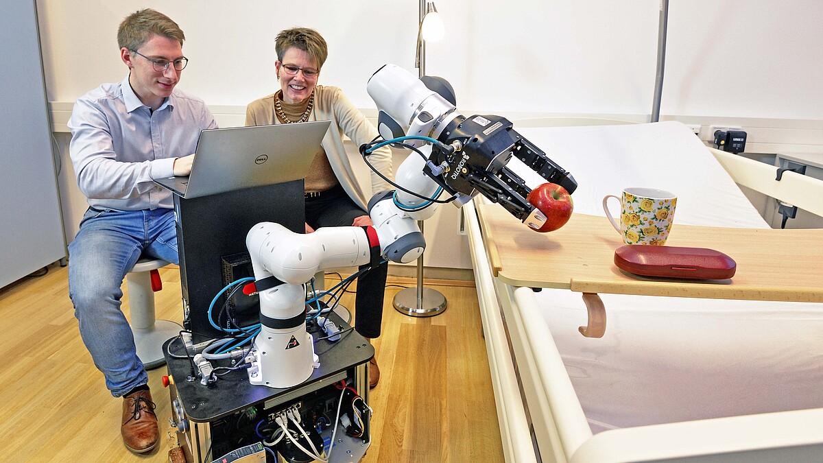 Zwei Personen mit Laptop sitzen neben einem Pflegebett. Ein Roboterarm greift einen Apfel.
