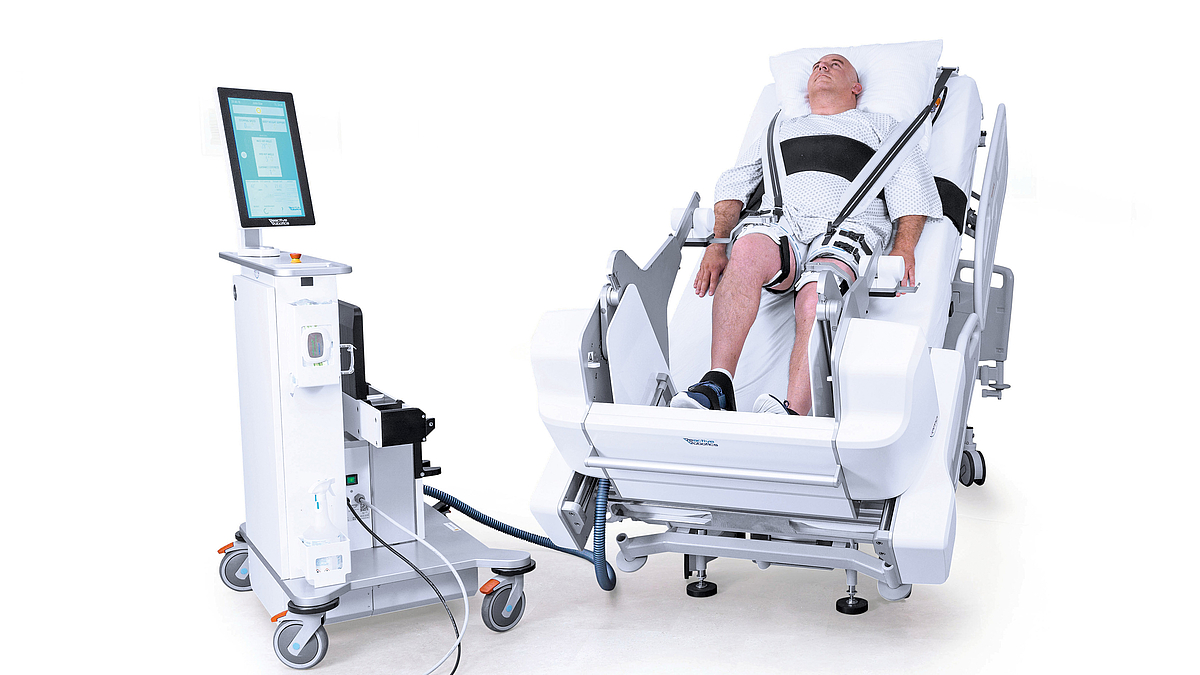 Ein Patient liegt in einem technisch aufgerüsteten Krankenhausbett. Daneben steht ein Rollcontainer mit medizintechnischer Ausrüstung.