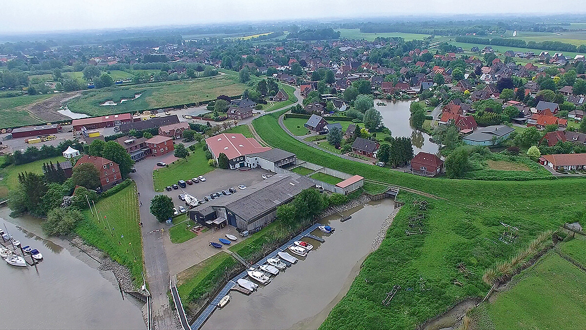 Pumpenhaus an einem Gewässer hinterm Deich mit Vor- und Hinterland, Siedlungen, Gewerbegebiet und Bootsanleger