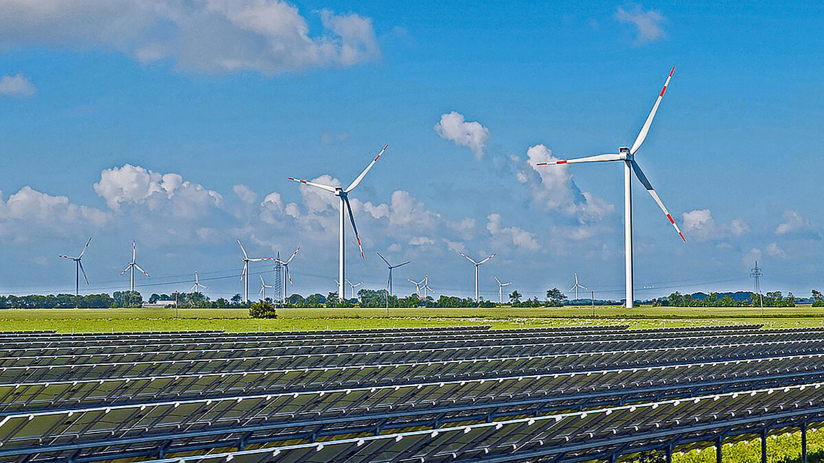 Vor einem Windpark stehen Reihen von Solaranlagen auf einer Wiese.