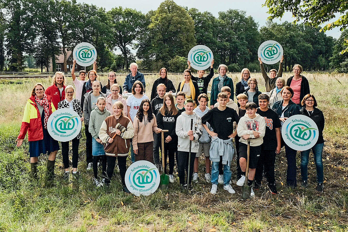 Eine Gruppe Jugendlicher und Erwachsener stehen mit Spaten auf einer von Bäumen gesäumten Wiese. Einige halten das Emblem „Schulwälder gegen Klimawandel“ hoch.