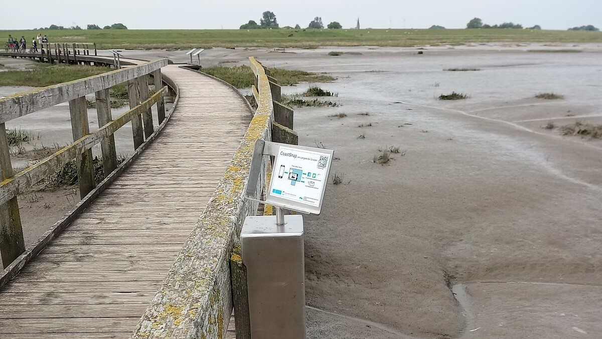 Ein Steg führt über den Wattboden zum Deich. Ein Schild fordert zum Fotografieren und Teilen auf.