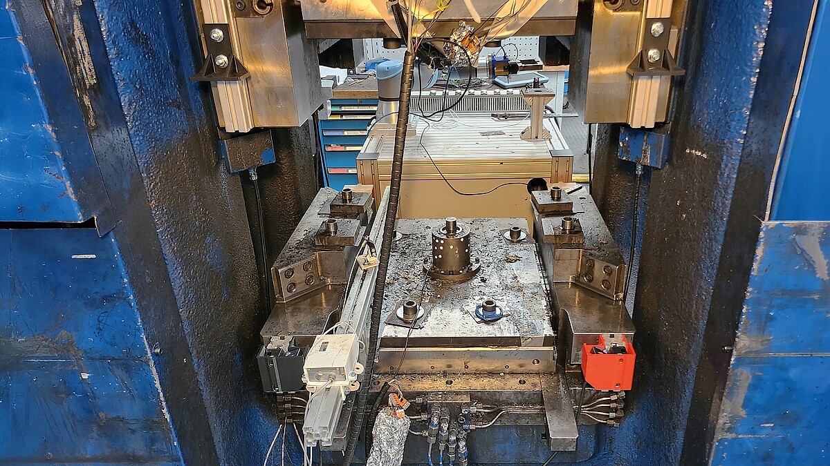 Eine geöffnete industrielle Presse mit einer Arbeitsplatte, auf der Metallteile und Halterungen montiert sind. Im Zentrum ist ein zylindrisches Werkzeug sichtbar. Oberhalb der Presse befinden sich elektrische Kabel und Sensoren. Im Hintergrund sind eine Arbeitsfläche, ein Roboterarm und Schubladen mit Werkzeugen zu erkennen.