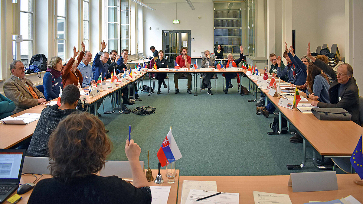 Eine Gruppe von Personen sitzt um ein Tisch-Karree mit Länder-Flaggen, einige heben die Hand zur Abstimmung.