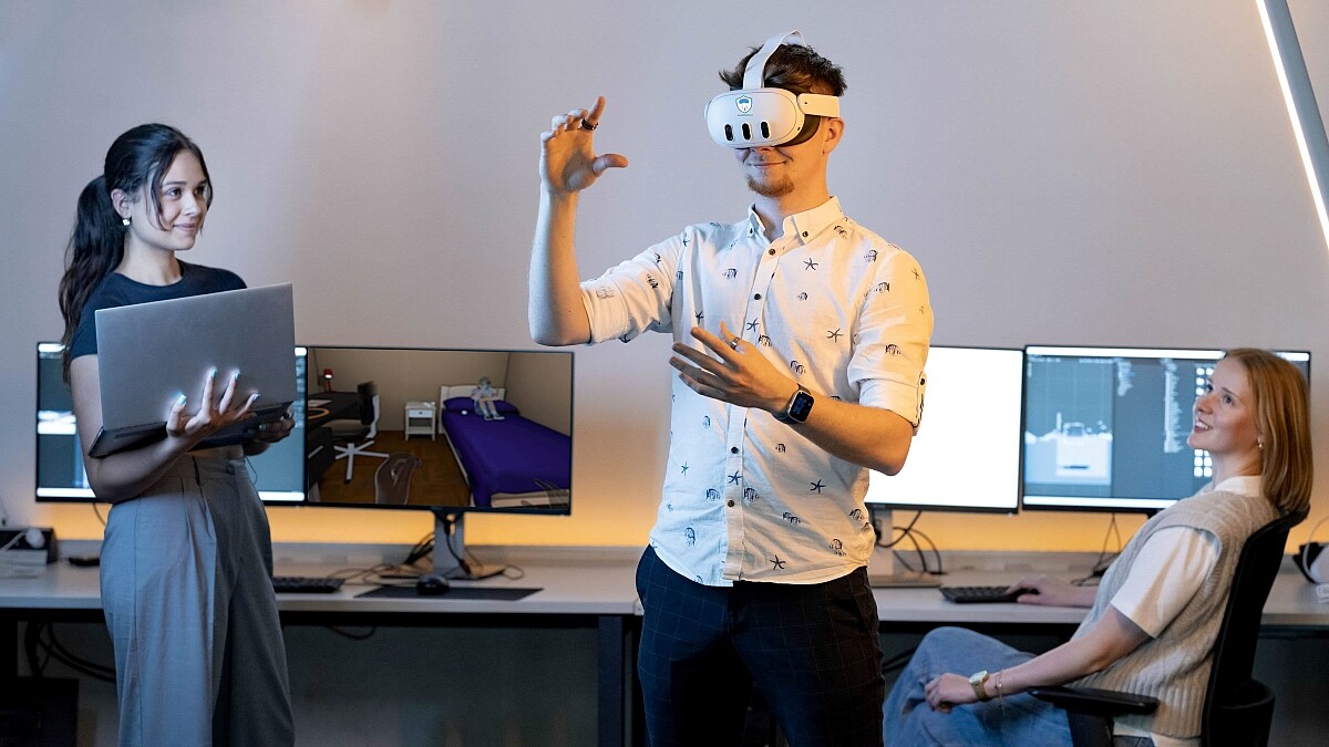 Ein Mann mit VR-Brille gestikuliert mit seinen Händen in der Luft. Zwei Frauen beobachten ihn. Eine hält einen Laptop im Arm, die andere sitzt an einem Bildschirm.