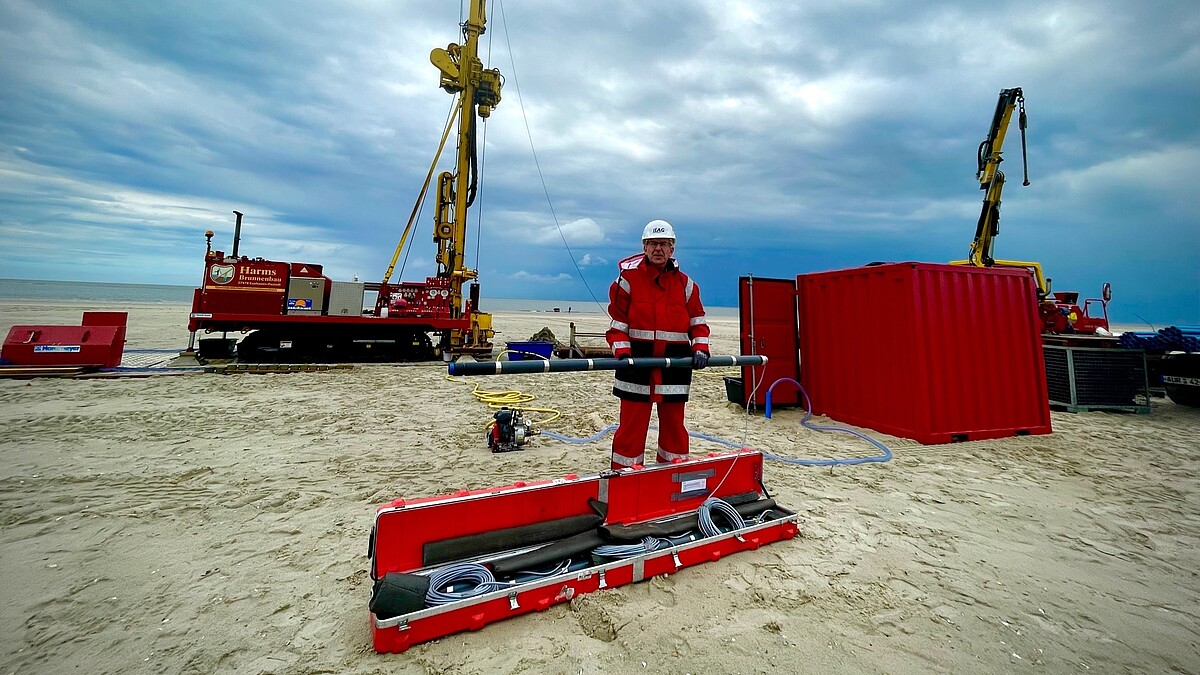 Am Strand stehen ein Bohrturm, Container und Kisten mit Material. Ein Mann mit Schutzkleidung und Helm steht davor und hält Bohrausrüstung in den Händen.