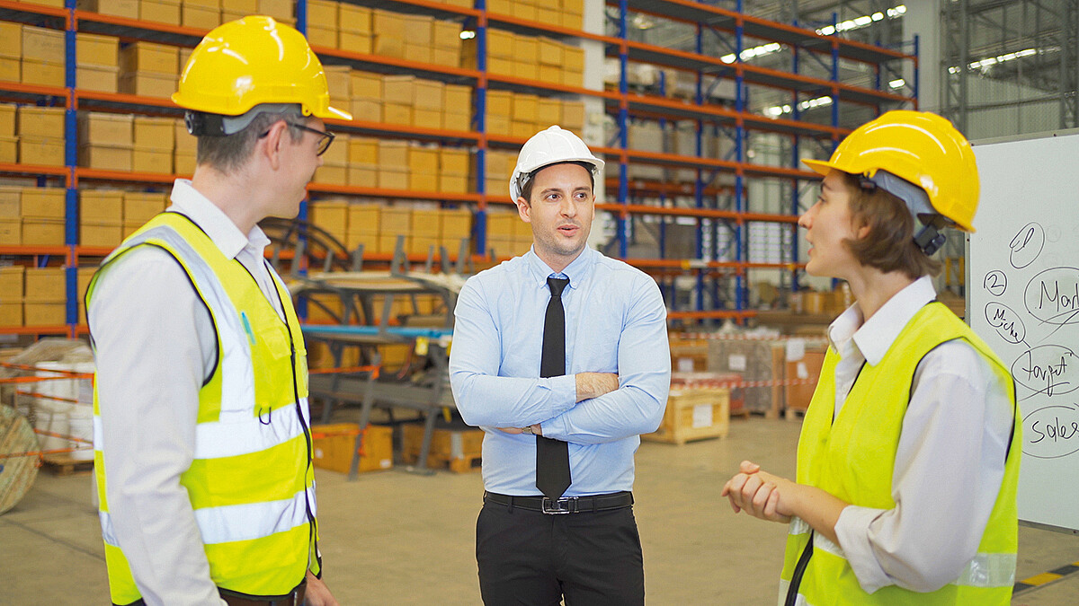 Drei Personen mit Business-Outfit beziehungsweise Warnwesten und Helmen besprechen sich in einem Warenlager.