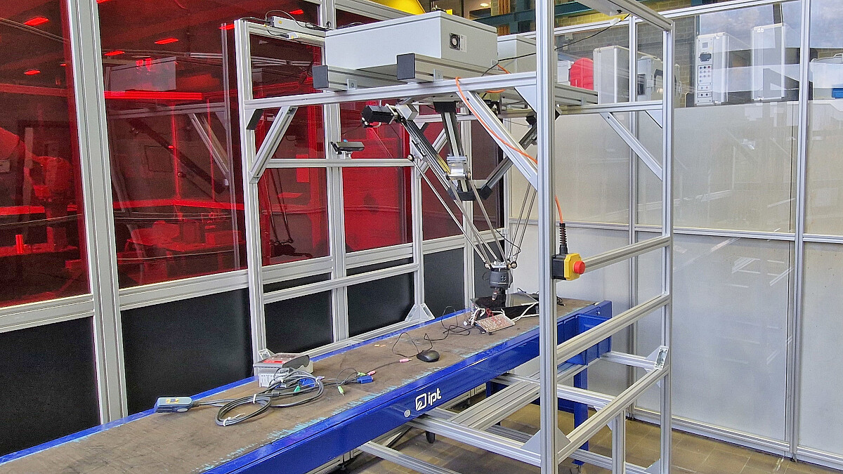 Über einem Förderband mit kleinen Elektrogeräten steht ein Gerüst mit Roboterarm, Kameras und Steuereinheiten.