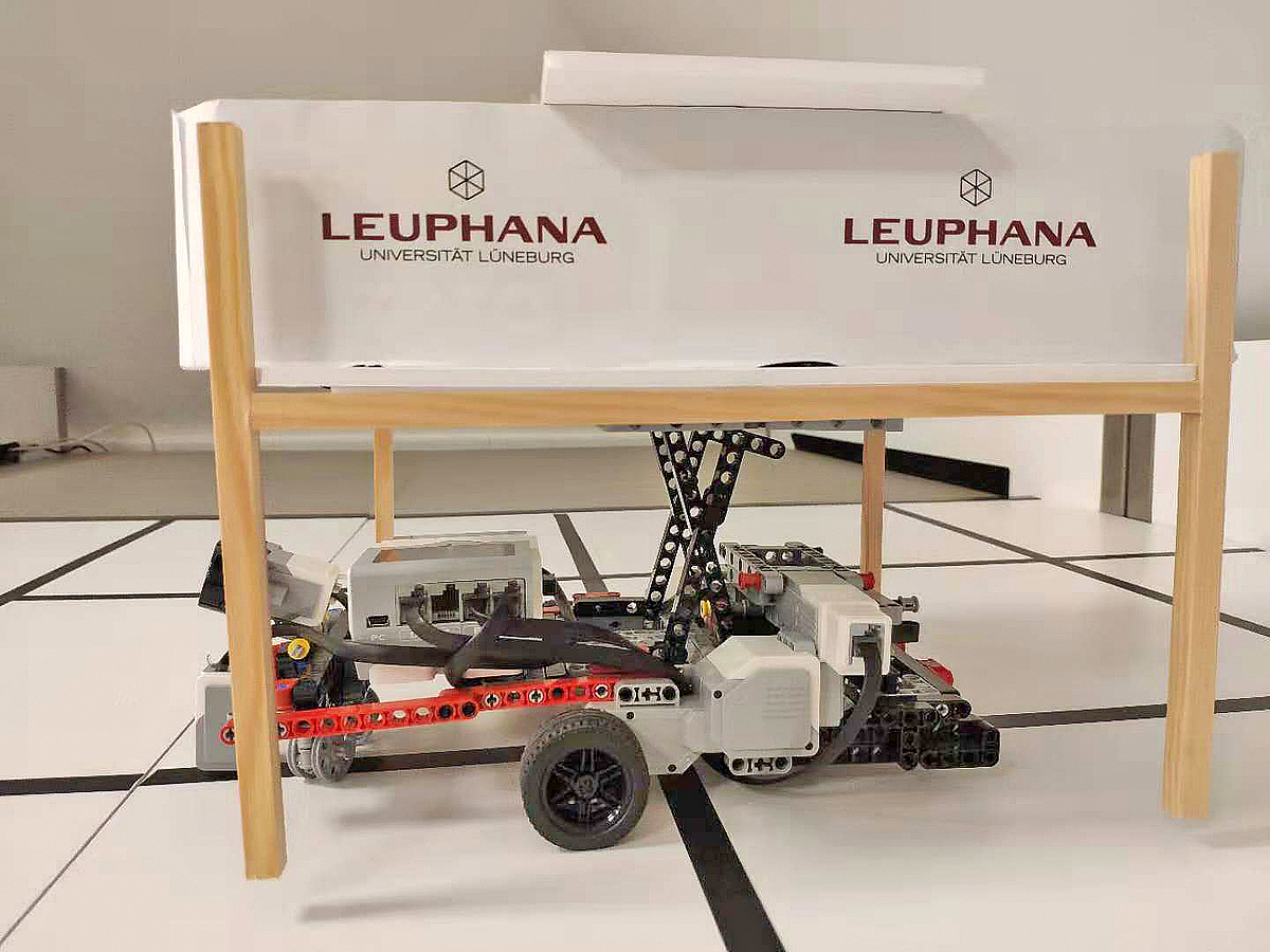 Ein Modellroboter hebt eine Transportbox auf Standbeinen über den Boden.