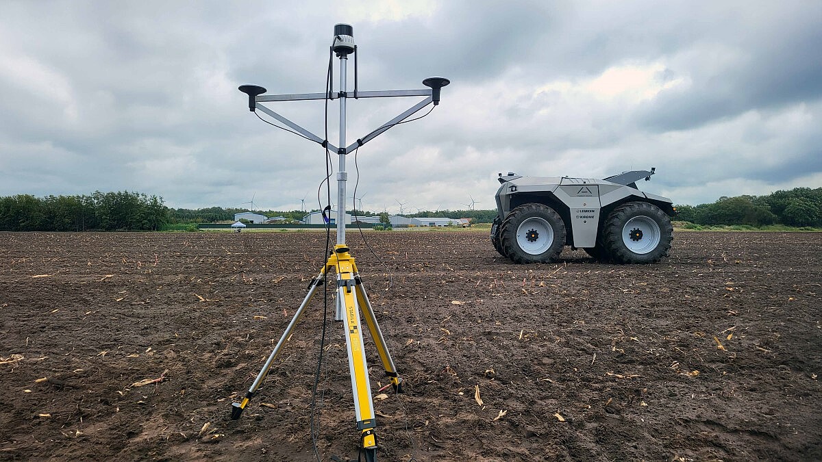 Das Foto zeigt eine autonome Zugeinheit mit dicken Rädern auf einem Acker. Im Vordergrund steht ein Vermessungs-Gerät.