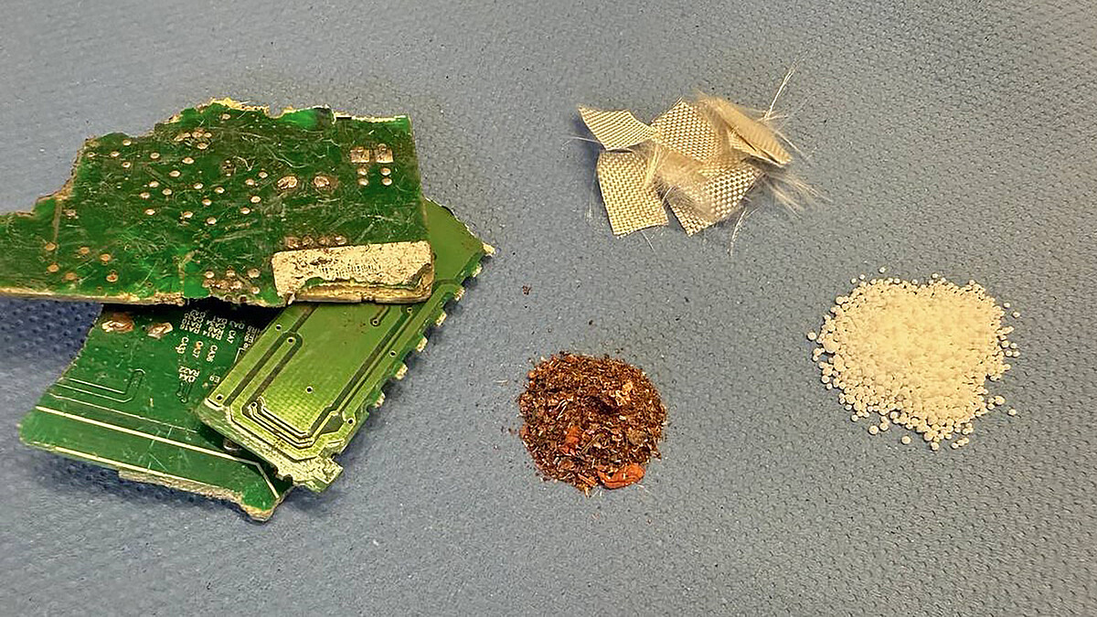Das Bild zeigt geschredderte Leiterplatten mit elektronischen Bauteilen und drei getrennte Häufchen aus Kupferspänen, Gewebeteilen aus Glasfasern und hellen Kügelchen.