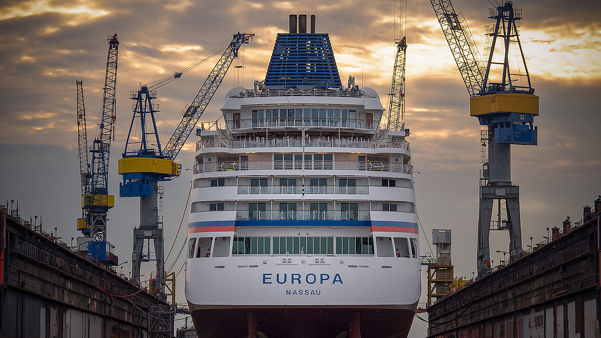 Mehrere Baukräne stehen um ein Kreuzfahrtschiff im Trockendock herum.