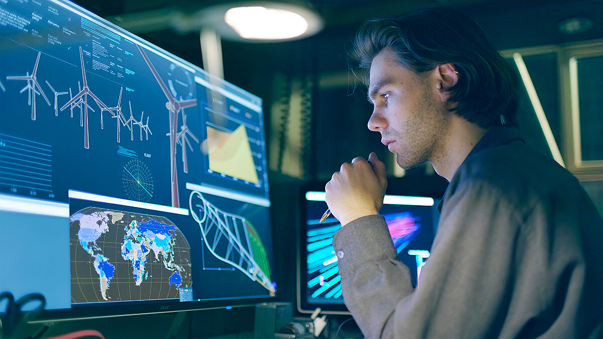 Eine Person beobachtet auf einem großen Monitor Daten zu Windkraftanlagen, eine globale Übersichtskarte sowie mathematische Modelle.