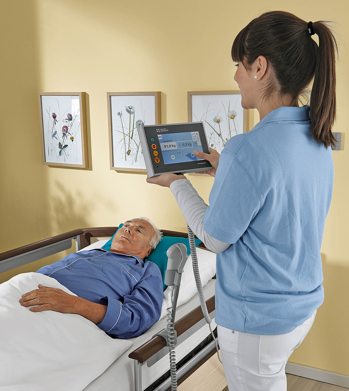 Eine Pflegerin hält ein digitales Tablet in der Hand. Das ist mit einem Bett verbunden, in dem ein Senior liegt.