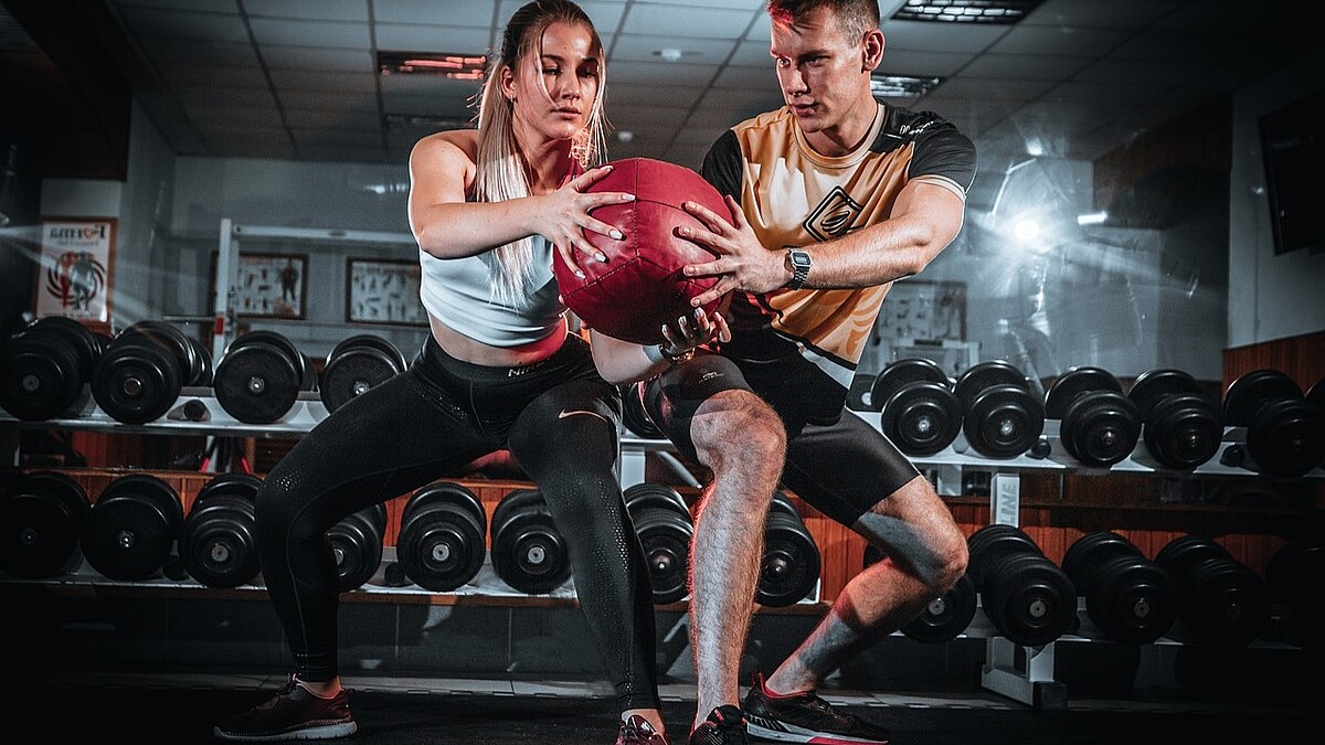 Eine Frau und ein Mann in Fitnesskleidung halten in der Kniebeuge gemeinsam einen Medizinball. Hinter ihnen lagern Hanteln.