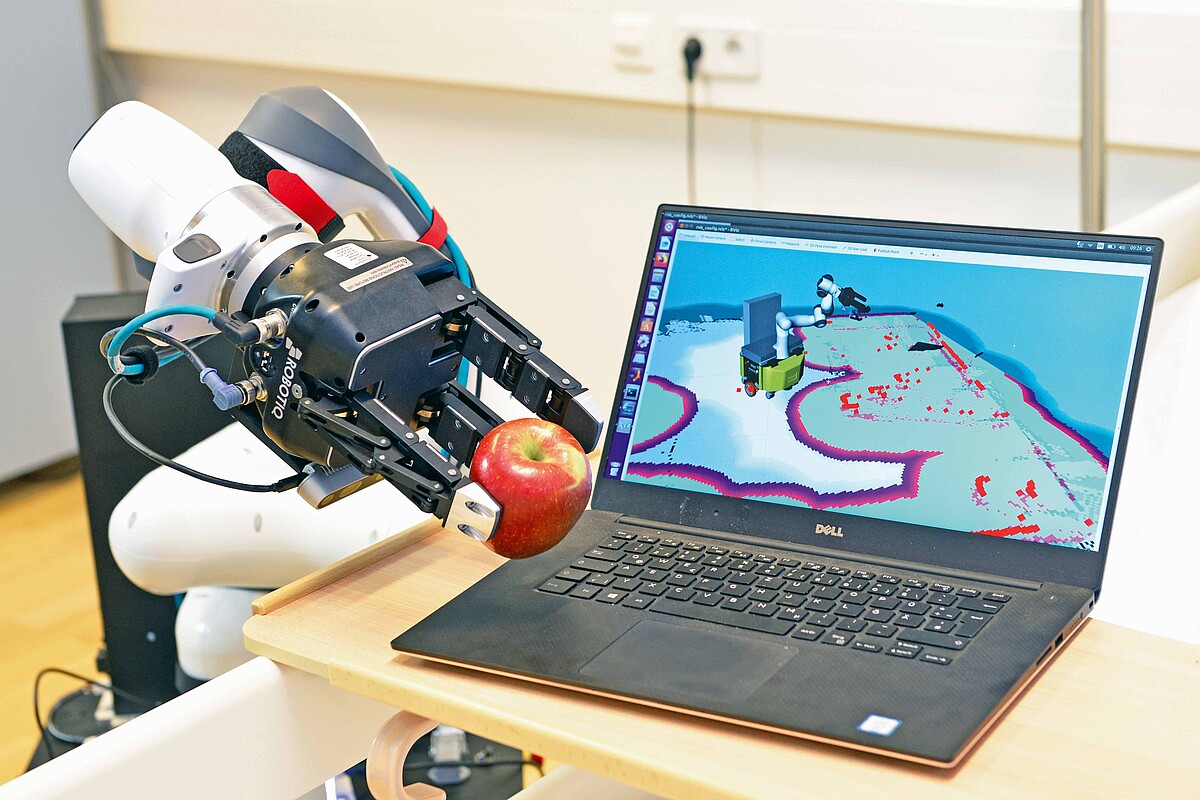 Ein Roboterarm legt einen Apfel auf einen Laptop. Auf dem Display ist der Vorgang digital abgebildet.