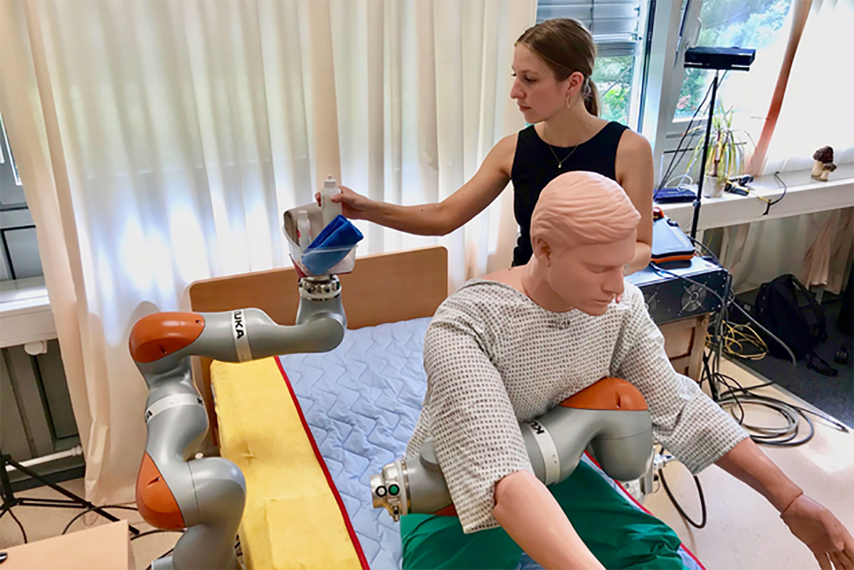 Das Roboter-Assistenzsystem bietet einer nach vorn geneigten Krankenpflegepuppe Unterstützung beim Sitzen. Hinter der Puppe steht eine Person, die nach dem Waschzeug auf dem Roboter greift