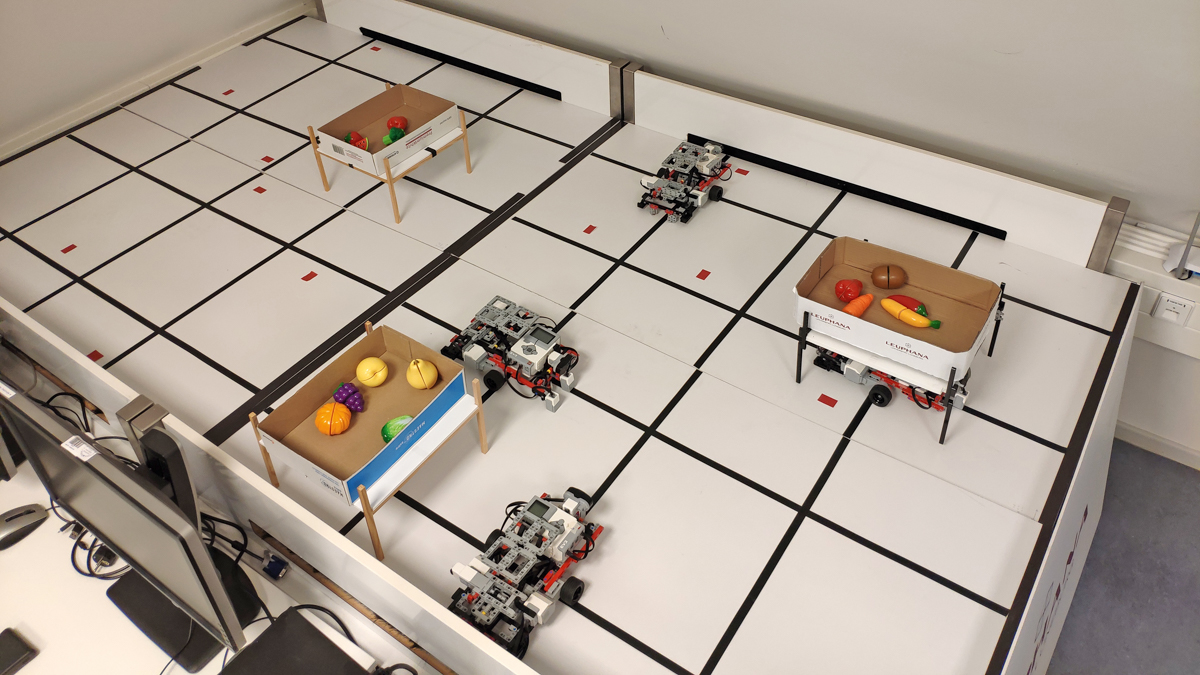 Modell-Roboter fahren auf einem mit Gitterlinien markierten Tisch, auf dem Transportboxen mit Spielzeug stehen.