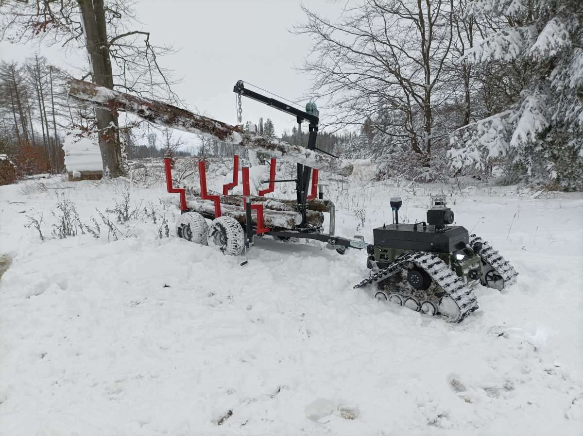 Ein kleines Kettenfahrzeug zieht einen Anhänger durch den Schnee. An einem Greifarm baumelt ein Baumstamm.