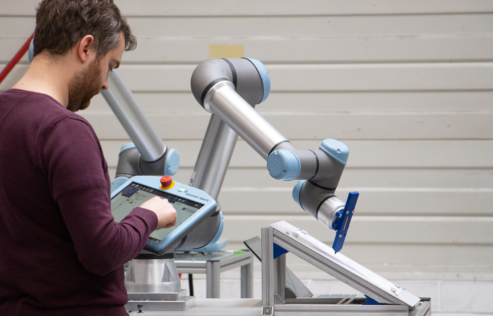 Ein Mann bedient ein Tablet. Neben ihm führt ein Roboterarm einen Stift über ein Zeichenblatt.