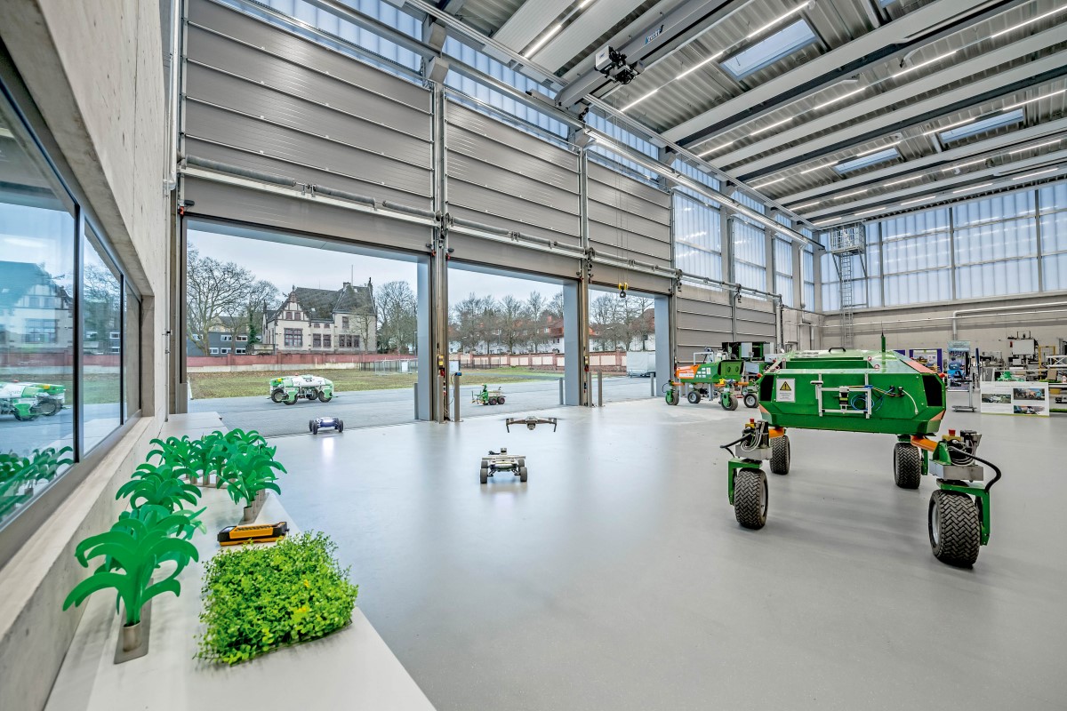 In einer Werkhalle befinden sich verschieden große Agarroboter auf Rädern sowie eine fliegende Drohne. Durch die geöffneten Rolltore ist das grüne Außenareal zu sehen.
