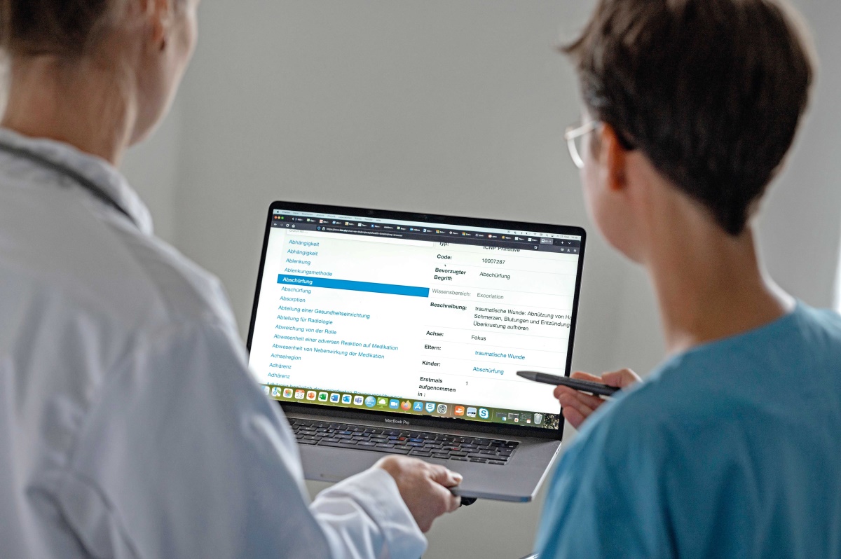 Zwei Personen schauen auf einen Laptop-Monitor. Dort sind Angaben zu "Abschürfung" und "traumatische Wunde" zu lesen.