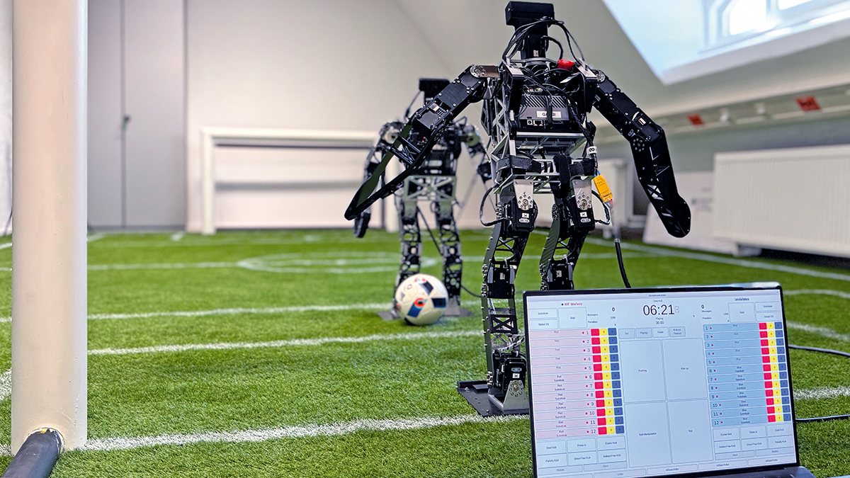 Zwei humanoide Roboter in Modellgröße stehen mit einem Fußball auf Kunstrasen. Ein Bildschirm zeigt Werte für die Spielerzustände, Verwarnungen und Spielstände an.
