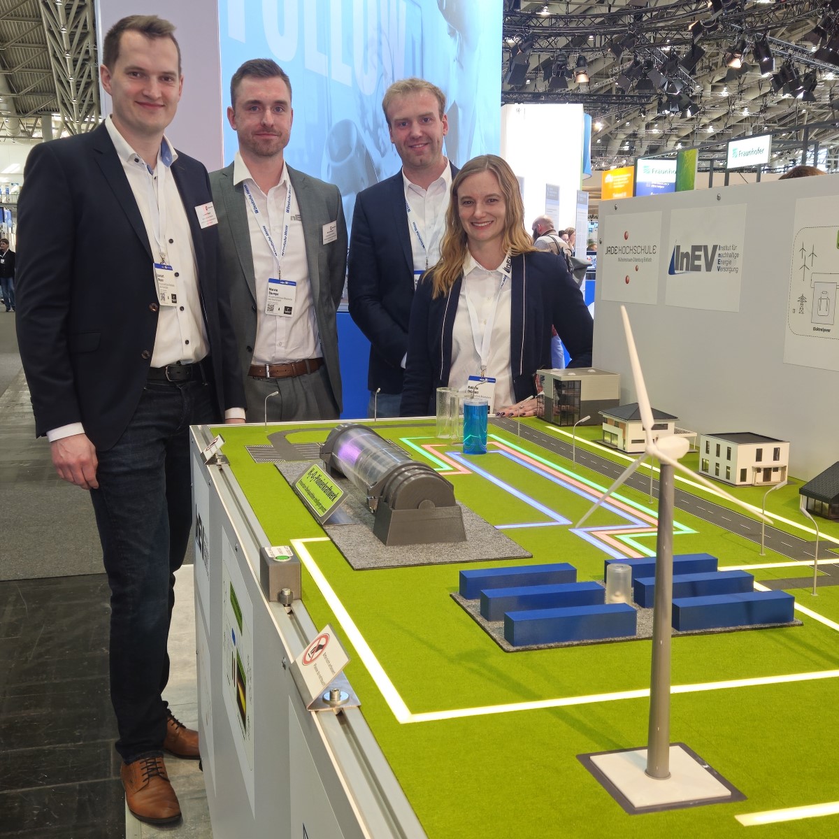 Eine Frau und drei Männer stehen auf einem Messestand hinter einem Modell. Auf einer Fläche mit Rasen und Straßen stehen Gebäude, Gas- und Wasserbehälter, Windkraftanlagen und eine Brennkammer.