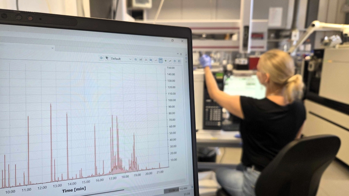 Eine Frau sitzt im Labor und bedient ein technisches Gerät. Im Vordergrund ist ein Monitor mit einem Diagramm zu sehen, dass über die Zeit verteilt unterschiedlich hohe Konzentrationen eines Stoffes darstellt.